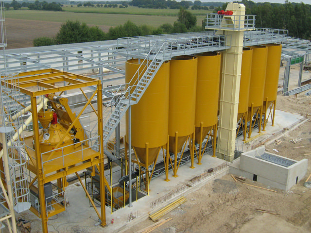 Bucket elevator pictured with 6 yellow silos and a concrete mixer.